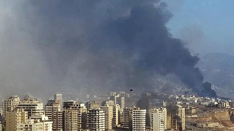 Aerial view of a coastal cityscape with dense smoke rising from multiple locations, suggesting conflict. In the foreground, a red Ferris wheel sits near the shore, juxtaposed against the chaos, conveying a tense atmosphere.