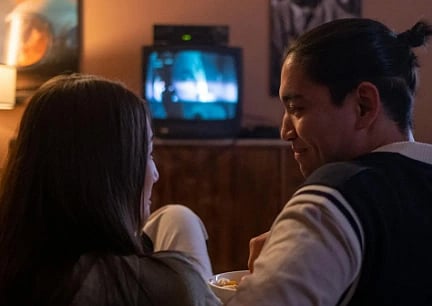 A couple sits on a couch in a cozy room, watching a movie on an old TV. They are smiling at each other, creating a warm and intimate atmosphere.