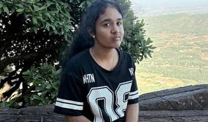 A girl with long hair sits on a rock, wearing a black sports jersey with the number 96. She is outdoors with green foliage and a scenic valley in the background.
