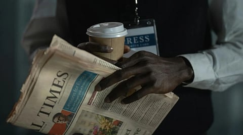 A person holds a coffee cup and newspaper. They're wearing a press badge and a shirt, suggesting a journalist's busy morning. The tone is professional.
