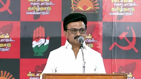 A man, DMK Chief MK Stalin, speaks at a podium against a backdrop featuring political symbols and Tamil text. The mood is formal and serious.
