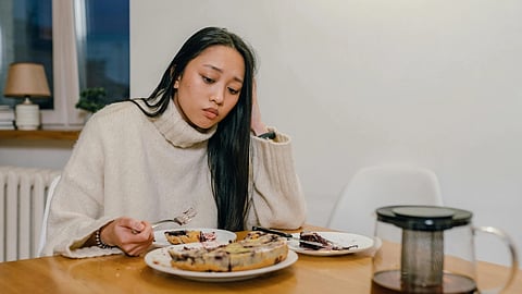 A woman in a cozy sweater sits at a table, looking contemplative. She has a fork in hand and a slice of pie on her plate, with a coffee pot nearby.