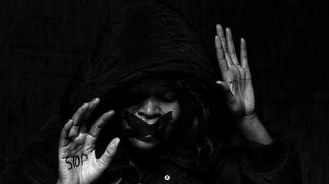 A person in a dark hooded jacket with tape over their mouth holds up their hands. The left palm has "Stop" written on it. The mood is serious and somber.