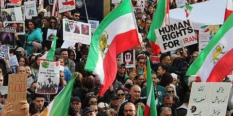 A large crowd of protesters hold Iranian flags and signs advocating for human rights in Iran. The atmosphere is energetic and united.