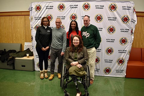 A diverse group of five people is smiling in front of a "City Year Chicago" backdrop. Sen. Tammy Duckworth is in a wheelchair is seated in the front. The mood is positive and inclusive.