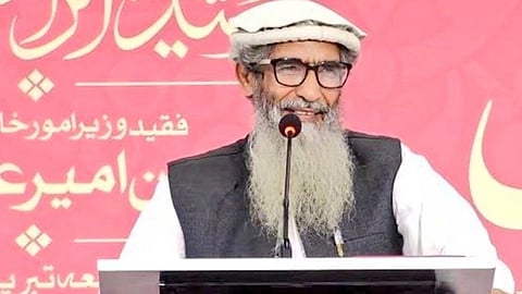 Elderly man with a long gray beard and glasses speaks at a podium. He wears a white cap and traditional clothing. The backdrop features text in Urdu.
