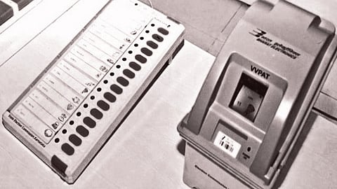 Electronic voting machine and VVPAT side by side on a white table. The voting machine has buttons next to names, while the VVPAT displays a paper slip.