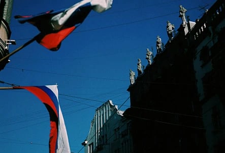 Flags with red, white, and blue stripes wave against a clear blue sky. Tall, ornate buildings with statues on top are partially visible.