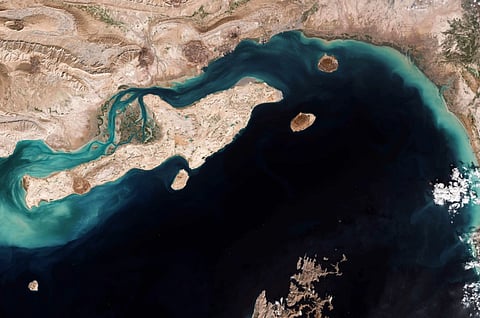 A satellite image (Strait of Hormuz) shows a large, irregularly shaped island surrounded by deep blue water. The island has rugged, sandy terrain with small vegetation.