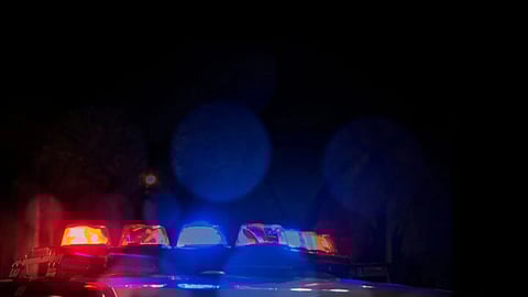 Police car lights flash red and blue in darkness, with "Police Line Do Not Cross" tape in foreground. Scene feels tense and urgent.
