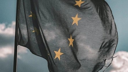 The image shows a tattered European Union flag waving against a cloudy sky. The flag's blue background contrasts with golden stars, conveying resilience.