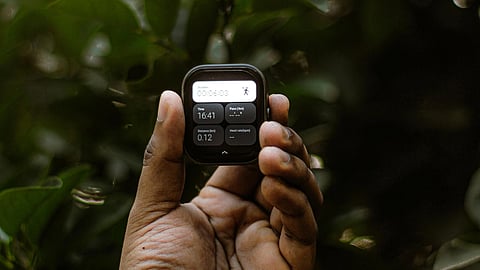 A hand wearing a black watch holds a small device with a screen displaying text. The background features lush green leaves, giving a serene outdoor feel.