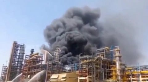 Thick black smoke billows from a large industrial complex, suggesting a fire. The scene conveys urgency, with firefighters spraying water in the foreground.