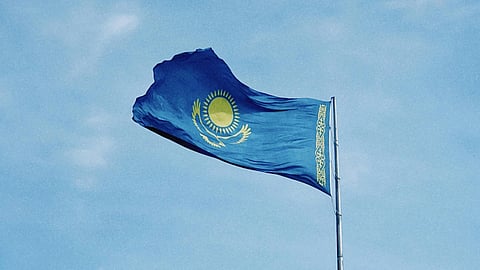 The flag of Kazakhstan waves against a clear blue sky. It features a gold sun with rays above a golden steppe eagle, with a vertical national pattern on the left.
