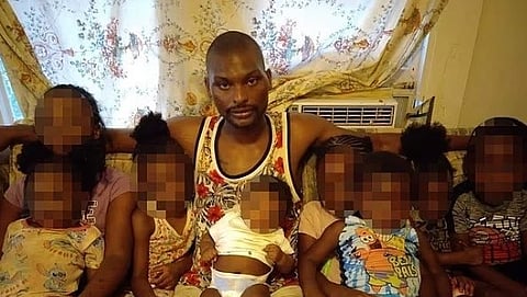 A man sits on a couch surrounded by several children. He wears a sleeveless shirt, and the children have colorful outfits. The room has a floral curtain and an air conditioner. The mood is warm and familial. Faces are pixelated for privacy.