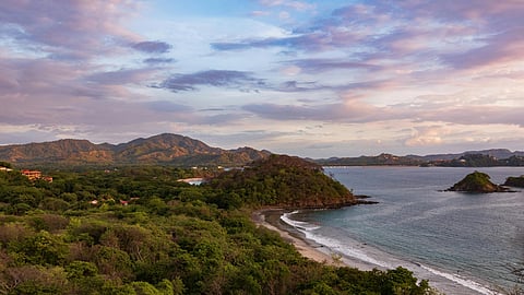 A serene coastal landscape at sunset, with lush green forested hills meeting a gently curving beach. Calm waves and colorful clouds enhance tranquility.