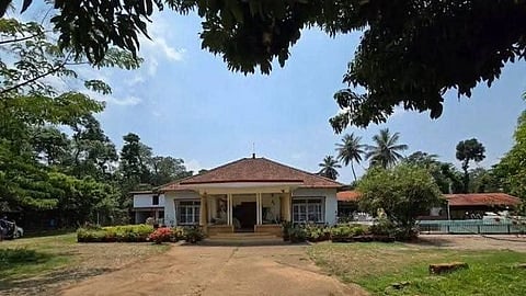 A charming single-story house with a red roof sits amidst lush greenery under a clear blue sky. Palm trees and a garden accentuate the serene setting.