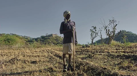 Elderly man stands in a dry field with a stick, wearing a turban and traditional attire. The landscape shows sparse trees and hills under a clear sky.