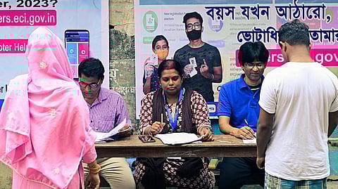 two people stand in front of three election officers with 2026 bengal election posters in the back