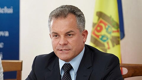 Vladimir Plahotniuc in a suit sits at a table with documents and glasses, in front of a blurred flag. He appears serious and engaged in a discussion.