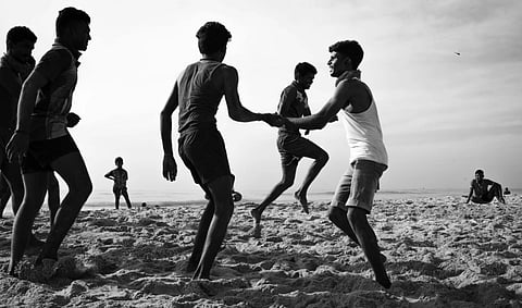 A black-and-white photo of men playing on a sandy beach. They are engaged in active movement, creating a lively and energetic atmosphere under a cloudy sky.