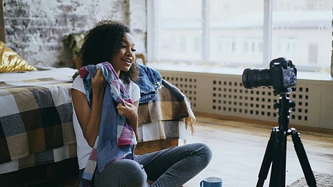 A person sits on a bedroom floor, holding colorful clothes, smiling at a camera on a tripod. The setting is cozy, with natural light from a large window.