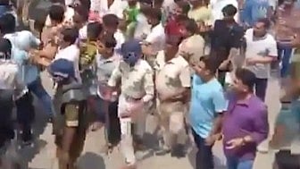 A large crowd of people, including police officers and civilians, walk together on a street in daylight. Some officers wear uniforms, creating an active and tense atmosphere.