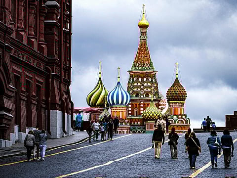 Red Square scene with people walking toward the colorful onion domes of St. Basil's Cathedral under a cloudy sky, flanked by historic red-brick buildings.