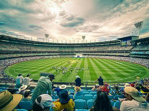 A vibrant cricket stadium filled with spectators. The lush green field hosts a match under a dramatic, cloudy sky. The atmosphere is lively and engaging.