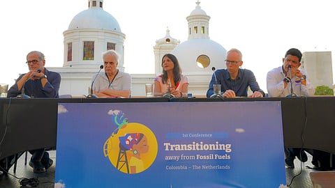 A panel of people sit behind a tale with a white building in the back and a board in front reading 'Transitioning Away from Fossil Fuels'