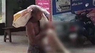 A shirtless man carries a large sack on his shoulder and holds a blurred object, walking on a street. Background includes a bench and a parked motorcycle.