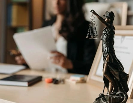 A statue of Lady Justice holding scales is in focus on a desk. In the background, a person in business attire is blurred, reviewing documents.