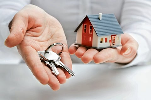 Two hands are shown; the left holds house keys and the right holds a miniature house model, symbolizing real estate and home ownership.