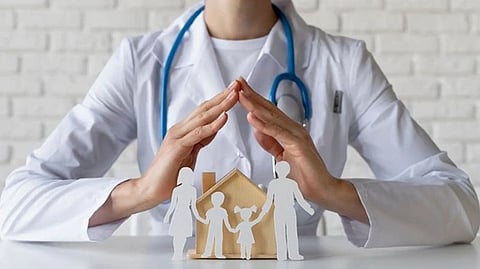 A doctor in a white coat and stethoscope cups their hands protectively over a wooden house model and paper cutout family, symbolizing healthcare support.