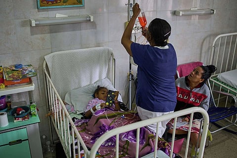 a child lies in a hospital bed while a nurse attaches an IV and a woman sits nearby