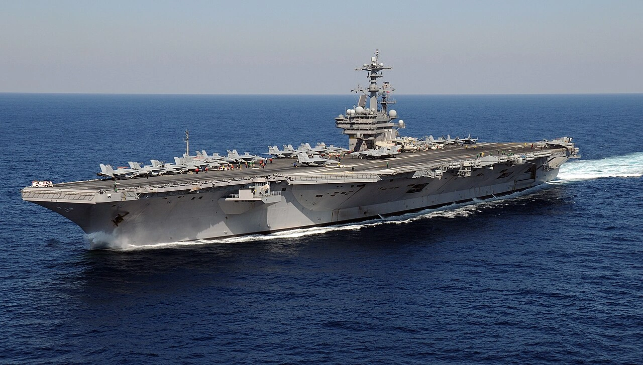 A large aircraft carrier sails on a calm blue ocean against a clear sky. Jets are lined up on deck, indicating military readiness and strength.