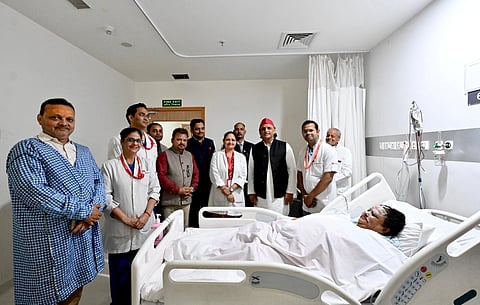 A group of people, including medical staff and visitors, stand around a patient lying in a hospital bed. The mood is supportive and attentive.
