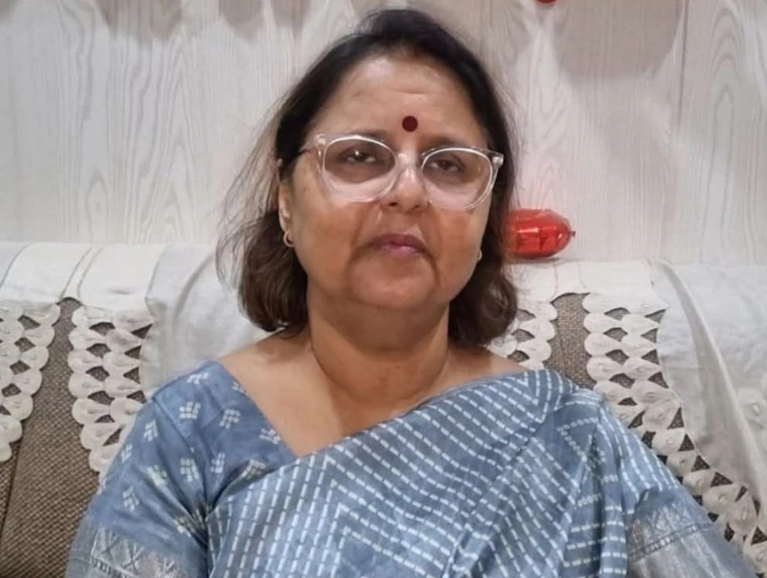A woman with glasses and a bindi sits on a sofa, wearing a gray patterned saree. The background features a light-colored wooden wall and cushions.