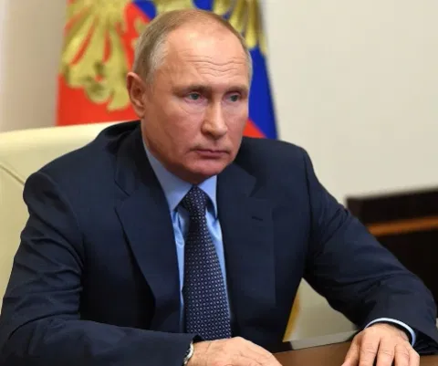 Image of Vladimir Putin sitting in front of a table with both his hands [placed at the table. he is wearing a dark blue suit.