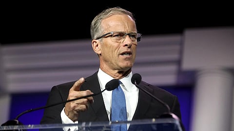 John Thune in a suit and tie speaks passionately at a podium with microphones against a backdrop with white columns, conveying a serious tone.