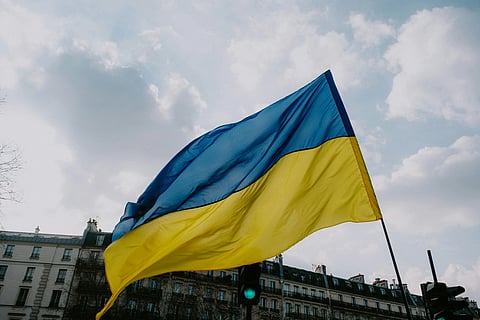 A Ukrainian flag waves prominently against a cloudy sky backdrop, with historic European-style architecture visible in the background, conveying a sense of national pride.