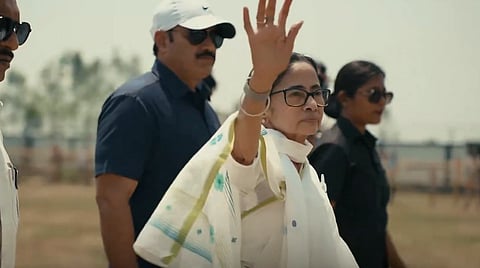 Image of Mamata banerjee in traditional attire waves confidently, flanked by security personnel in sunglasses. The setting is outdoors on a sunny day, conveying determination.