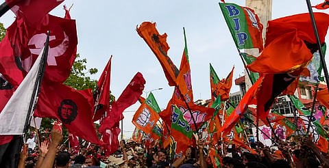 Crowd waving red and orange political flags with symbols and faces, set against a backdrop of trees and a partially cloudy sky, creating an energetic atmosphere.