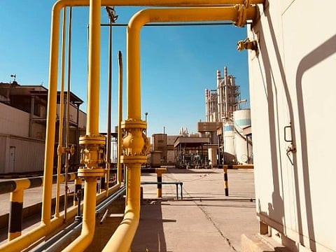 Yellow industrial pipes run along a factory wall under a clear blue sky, leading towards a distant refinery, conveying a sense of industry and efficiency.