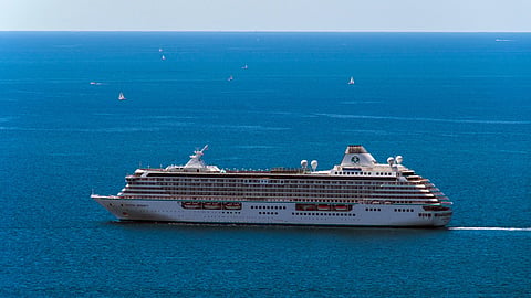 A large white cruise ship sails across a vast, calm ocean under a clear blue sky. Smaller sailboats are scattered across the distant horizon.