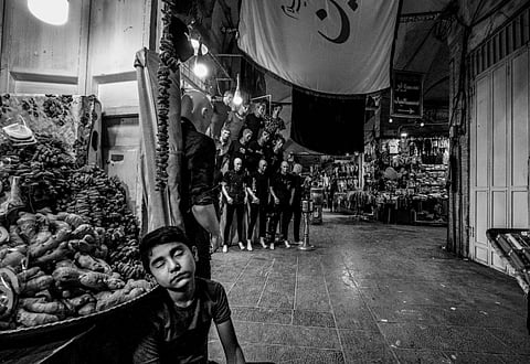 Black and white photo of a market scene. A young boy is asleep against a wall, surrounded by mannequins and produce, creating a serene yet busy atmosphere.