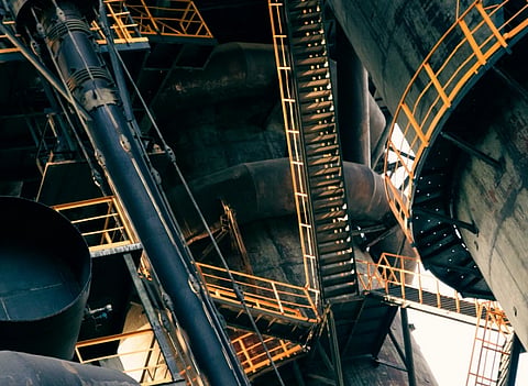 Industrial structure with interconnected steel pipes and orange-rimmed staircases. The dimly lit scene conveys a complex and mechanical tone.