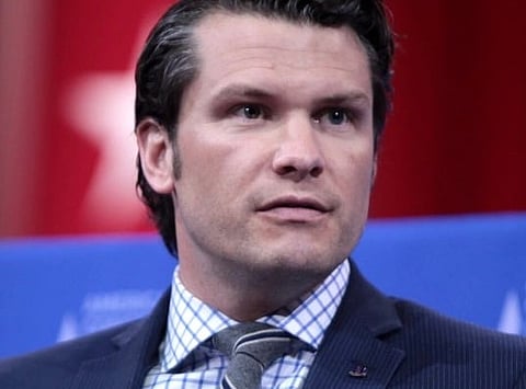 A man with a serious expression wearing a gray suit, striped tie, and checkered shirt. He is speaking into a microphone against a red and blue backdrop.