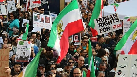 A large crowd of protesters hold Iranian flags and signs advocating for human rights in Iran. The atmosphere is energetic and united.