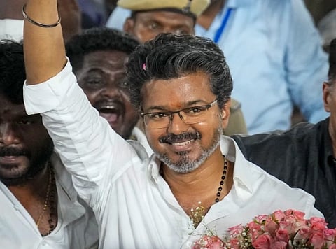 Image of Thalapathy Vijay in a white shirt raises his arm, holding a bouquet of pink roses. Surrounded by cheering crowd, conveying happiness and celebration.